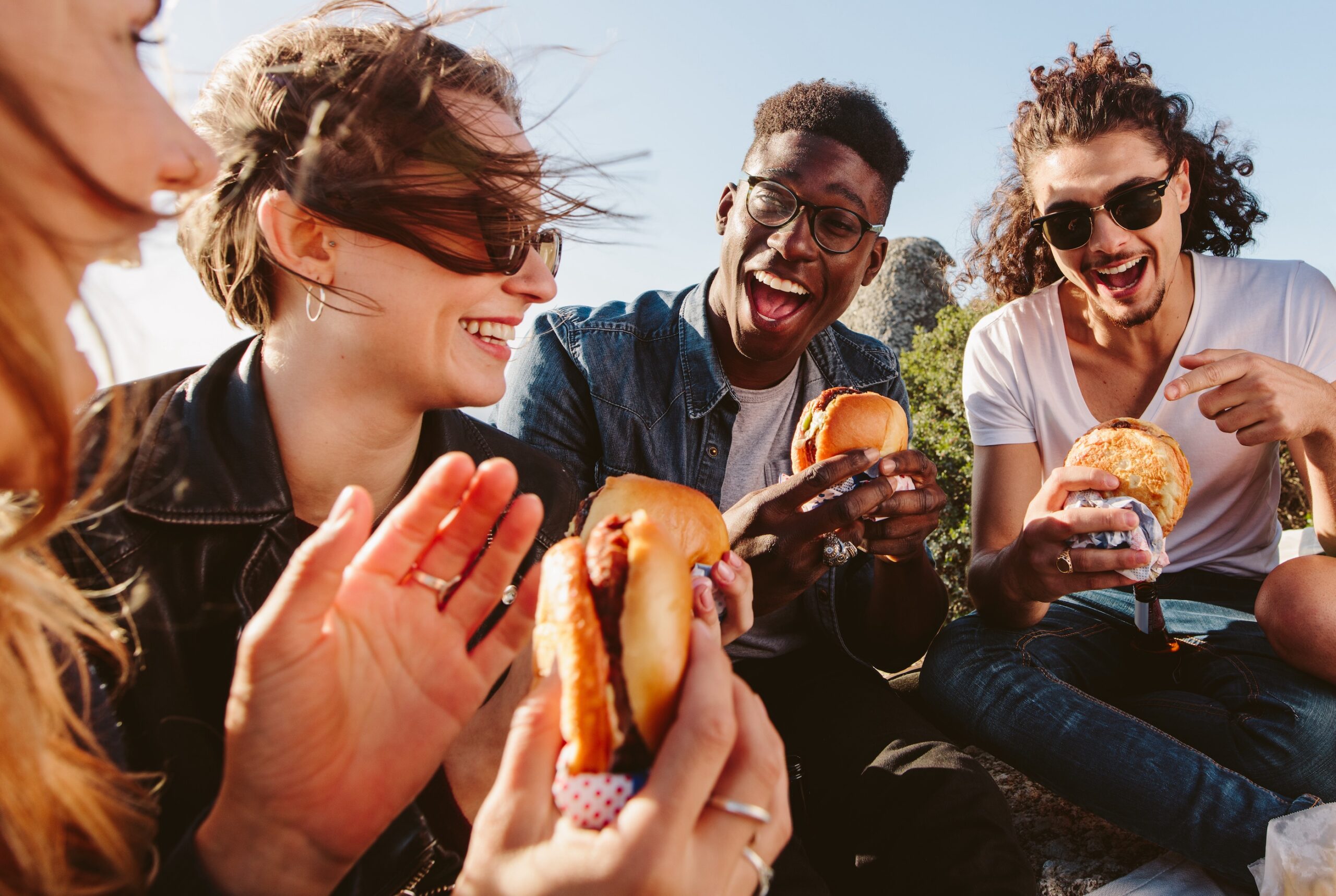 Diverse group of young friends laughing and enjoying burgers together outdoors on a sunny day scaled