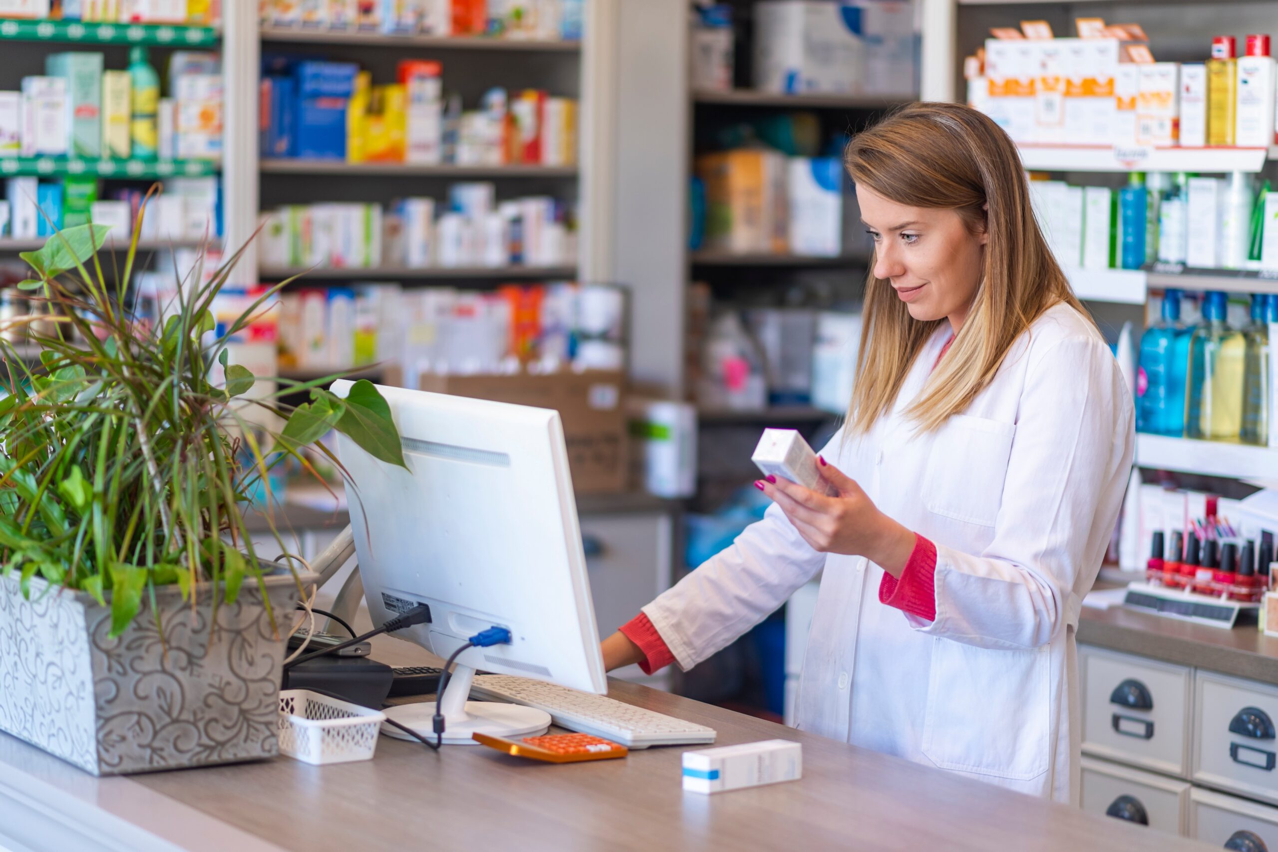 Female pharmacist in a white lab coat examining a medication box at a pharmacy counter behind a computer monitor scaled
